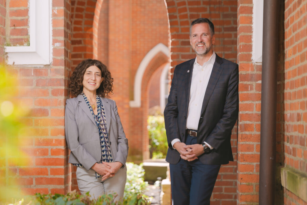 Lori and John CCG, brick building, two people standing side by side, leaning, grey suit, dark grey suit, button up shirt