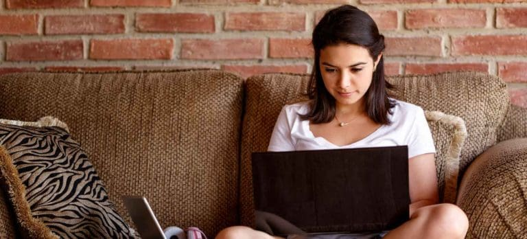 Woman sitting on a brown sofa with a laptop on her lap, focused on the screen in a living room with a brick wall behind her.