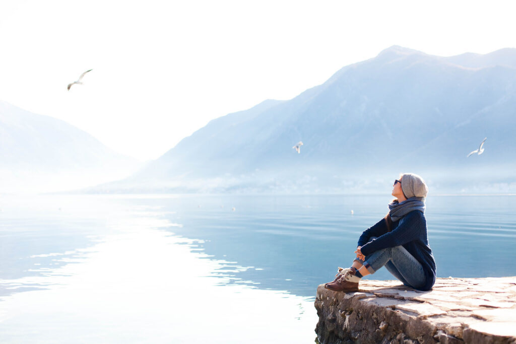 Young woman relaxing at winter sea beach Traveler resting by blue mountain | Antioch University Person in a beanie and scarf sits on a rocky ledge by a calm lake, with mountains in the background and seagulls flying above.
