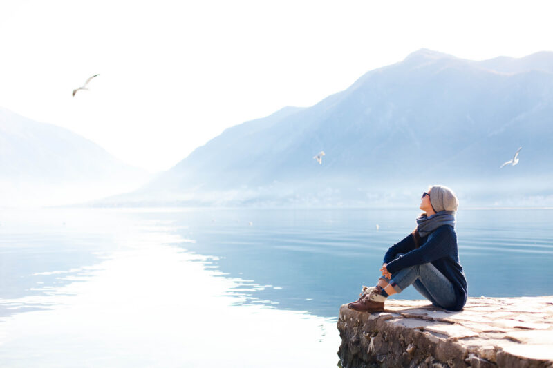 Person in a beanie and scarf sits on a rocky ledge by a calm lake, with mountains in the background and seagulls flying above.