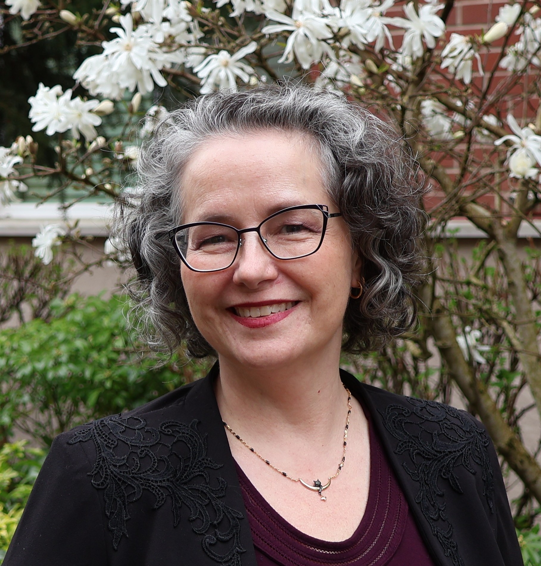 Smiling gray haired woman wearing glasses outdoors with white blossoms in the background
