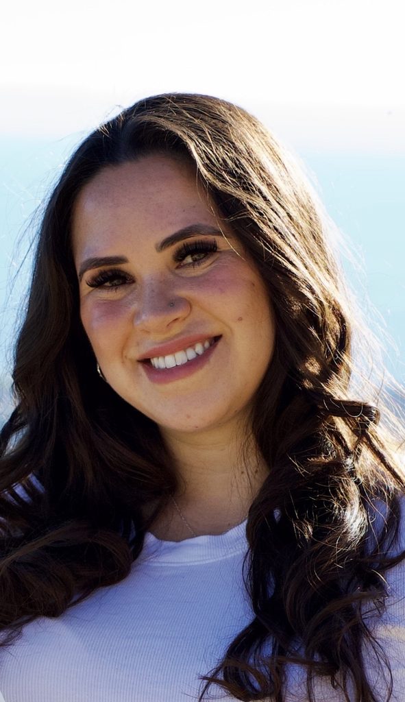 portrait of a woman with long wavy brown hair smiling in a white shirt by the ocean