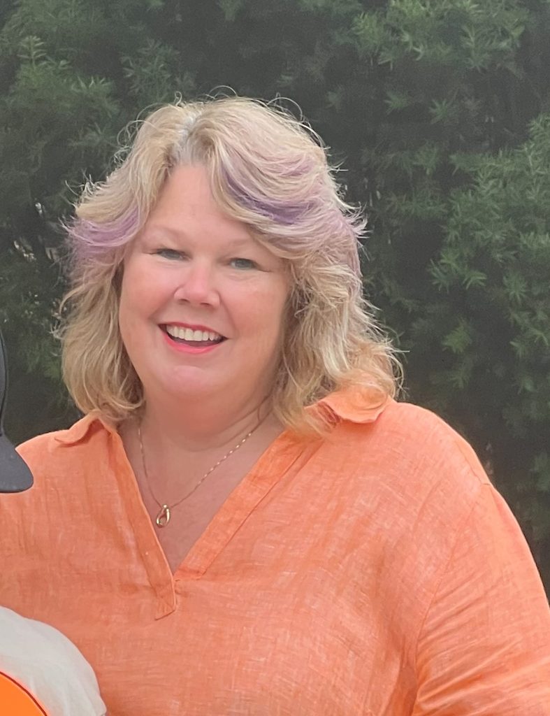 portrait of a woman with blonde hair in a bright orange shirt in front of bushes smiling