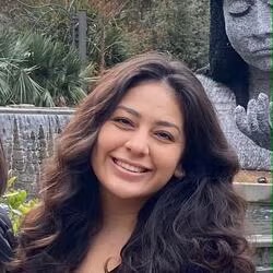 a smiling woman with long wavy brown hair posing in front of a waterfall and stone statue of a woman