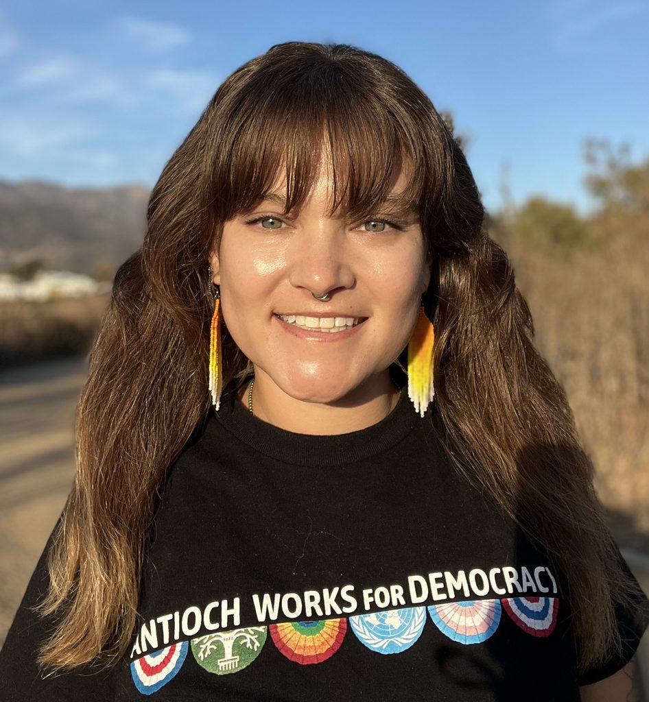 portrait of a woman with long brown hair smiling woman in a black Antioch University shirt a woman with bright orange earrings